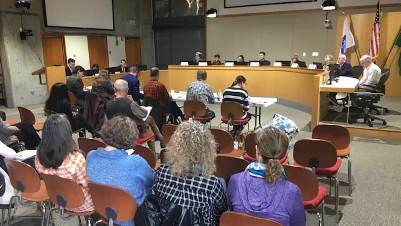 Photo from the back of an audience at a local council meeting; councilors sitting begind a raised counter at the front.