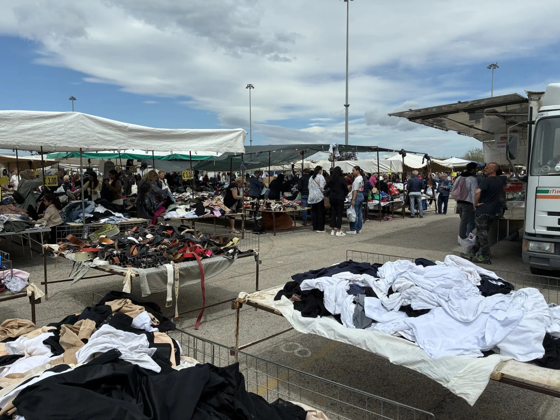Mercato settimanale popolare con bancarelle di abiti e oggetti usati sotto un cielo mediterraneo
