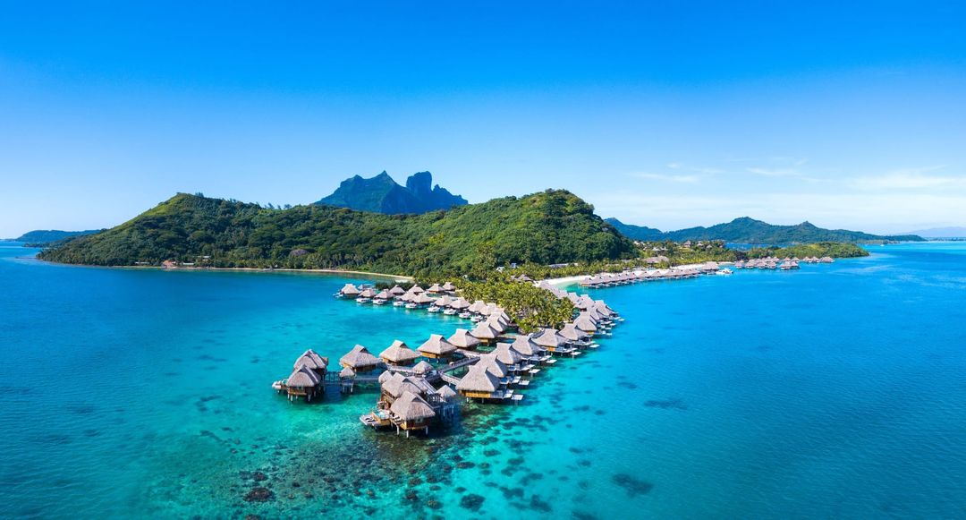 Conrad Bora Bora Nui from the air: a curve of overwater villas over a turquoise lagoon, Mount Otemanu behind