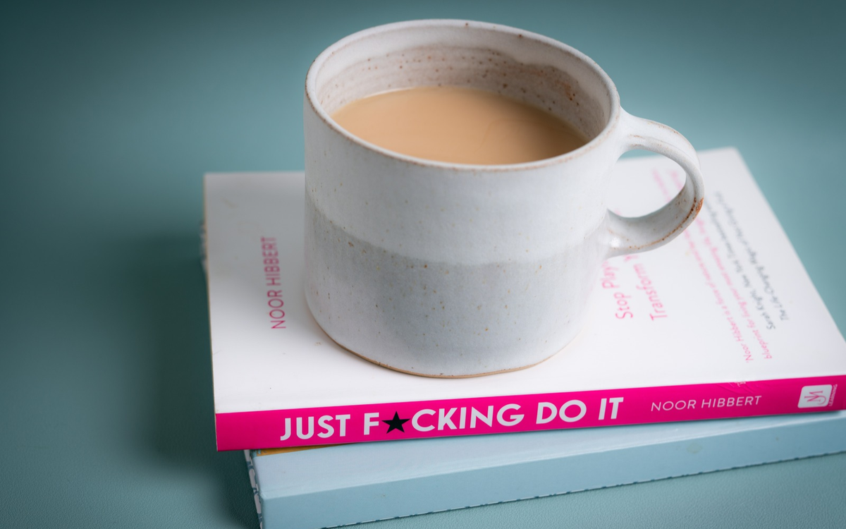 A mug of tea on a stack of books, the top one a pink-spined copy of Just F*cking Do It by Noor Hibbert