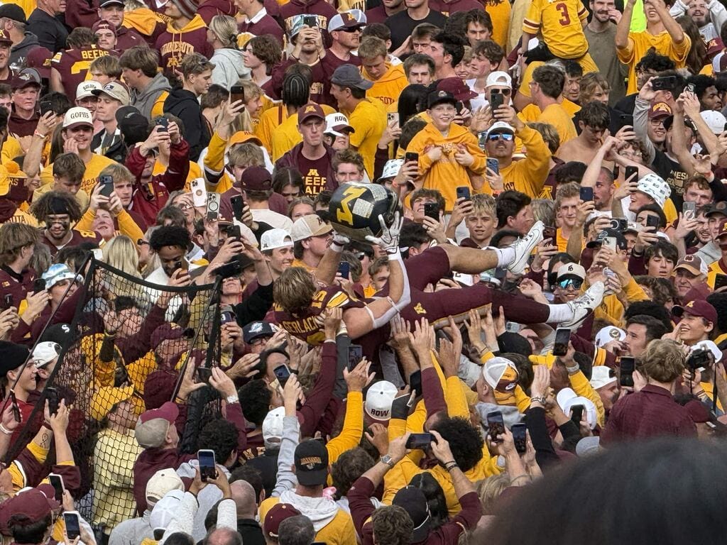 Crowd rushing the field with a Minnesota player riding the way, while holding the Little Brown Jug trophy.