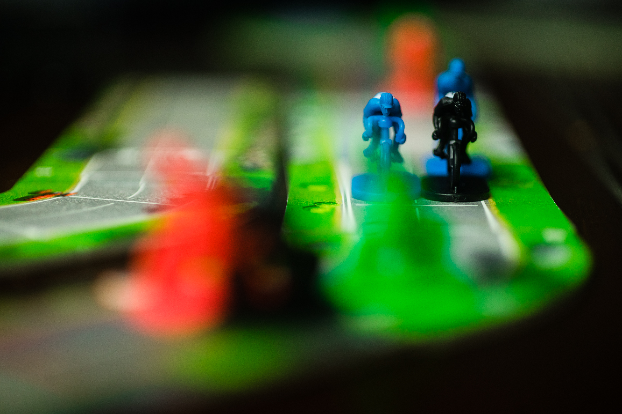 The board game Flamme Rouge, with two racing miniatures in view. There are racers in the foreground out of focus.