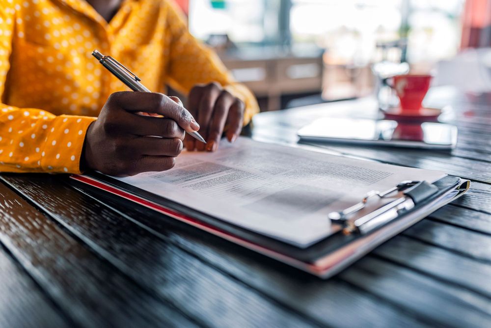 Person with dark skin wearing a yellow sweater filling out a survey on a clipboard