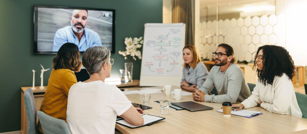Diverse team sitting at conference table with a member calling in on a video call