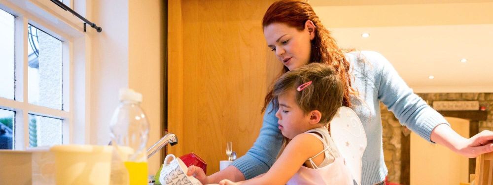 Little boy stands at the kitchen sink washing up. He is concentrating as a woman stands at the side to help.