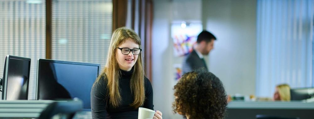 Two colleagues talk while holding cups of coffee. One sits in a desk chair, the other sits on the desk.