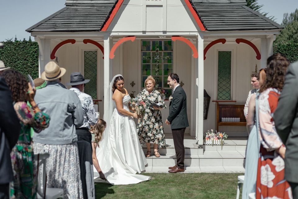 A bride and groom stand before your celebrant Ana Connor, smiling as the bridesmaid arranges the bridal gown.
