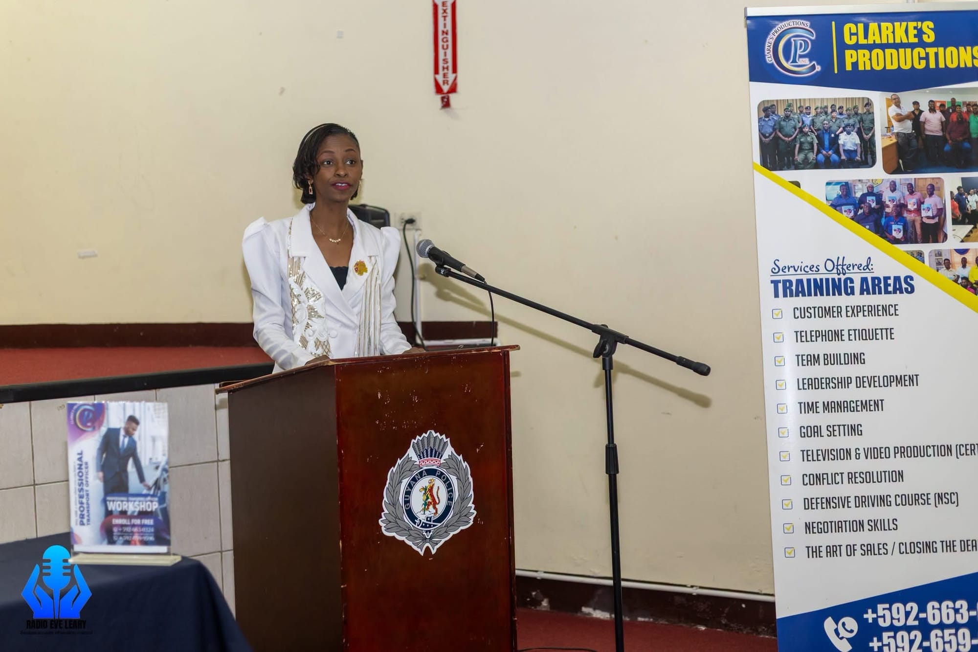 Ms. Carlyne Stewart, Chief Experience Officer of Clarke's Productions Inc., delivering remarks. The banner beside her lists the company's training areas, including the National Safety Council defensive driving course.