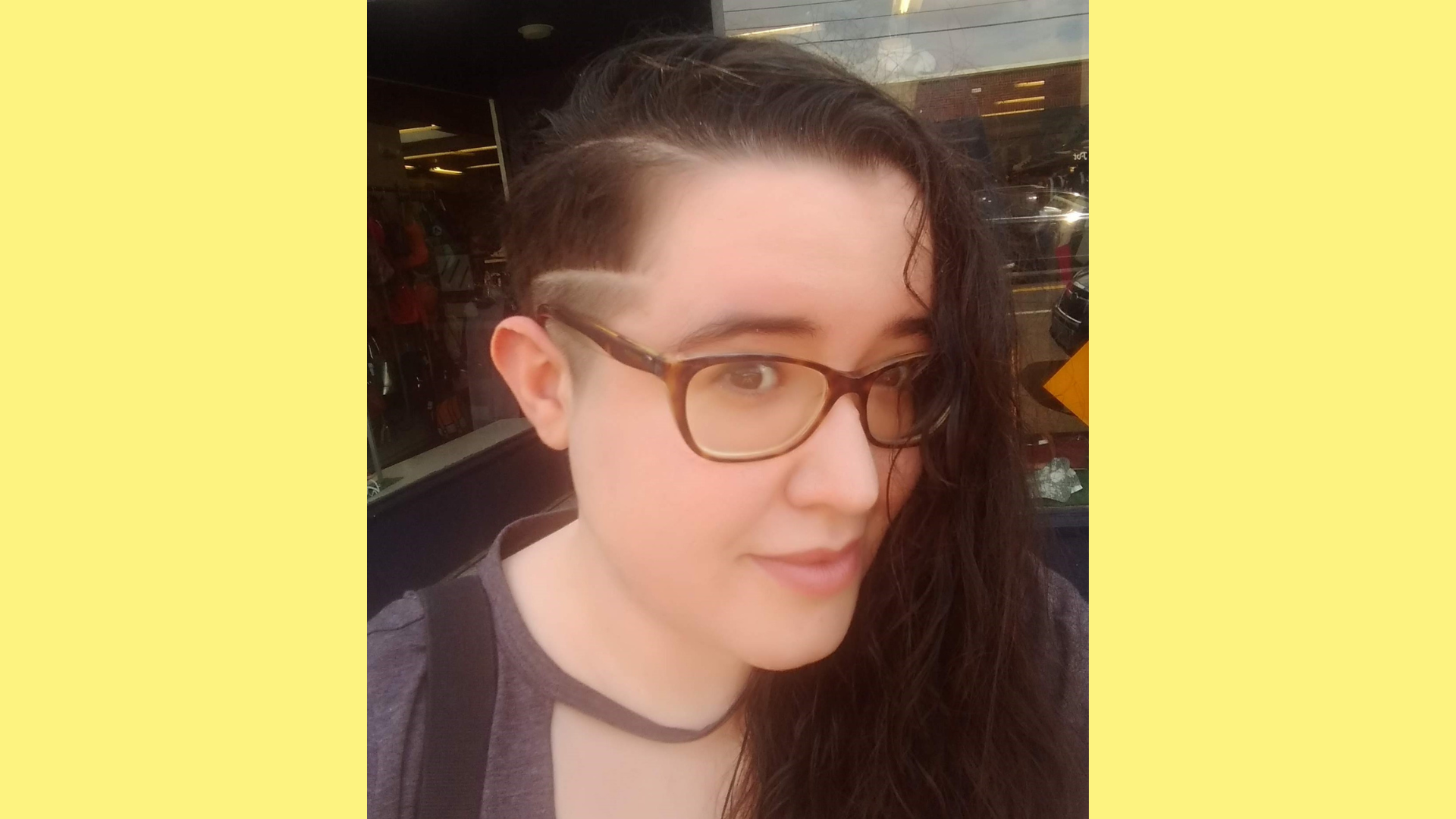 Ely smiling softly in front of a clothing store; their hair is cast over one side, revealing an undercut.