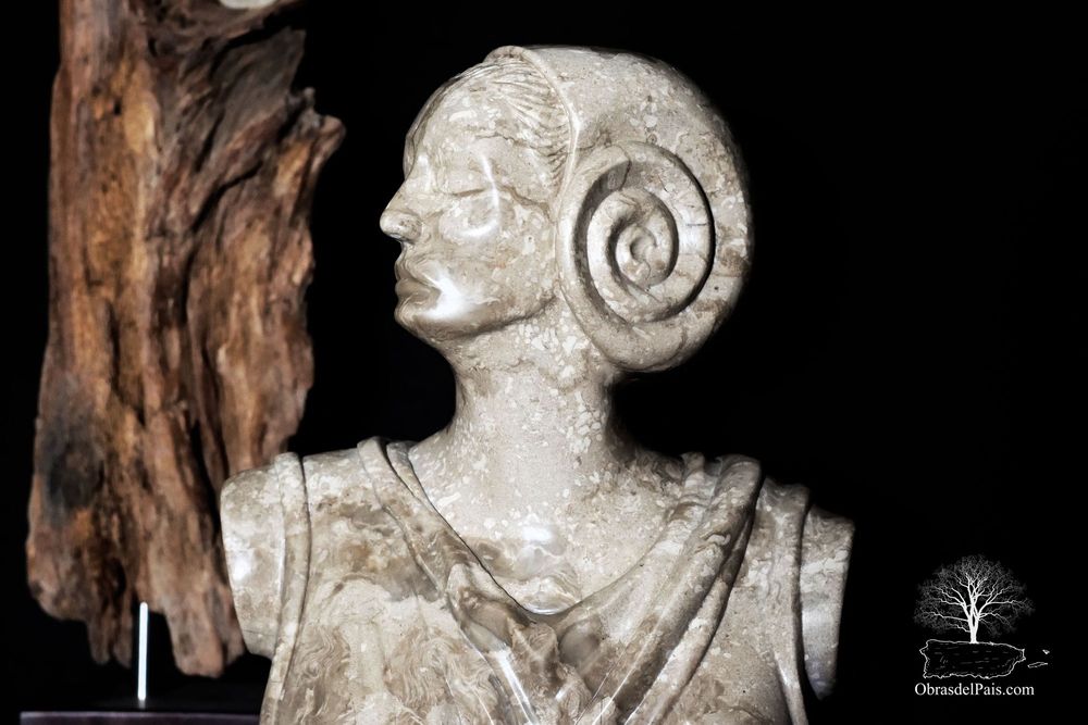 Busto femenino tallado en piedra clara, con una pieza de madera visible en el fondo.