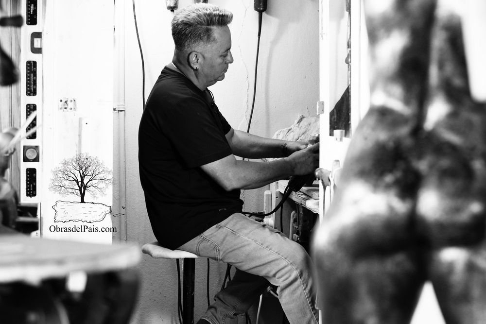 José Antonio Rodríguez sentado en su taller trabajando una pieza, con una escultura desenfocada en primer plano.