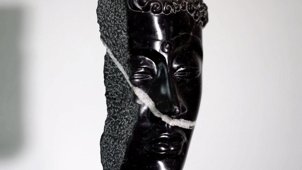 Escultura de un rostro en piedra negra con acabado pulido, borde rugoso y una veta clara que cruza la pieza.