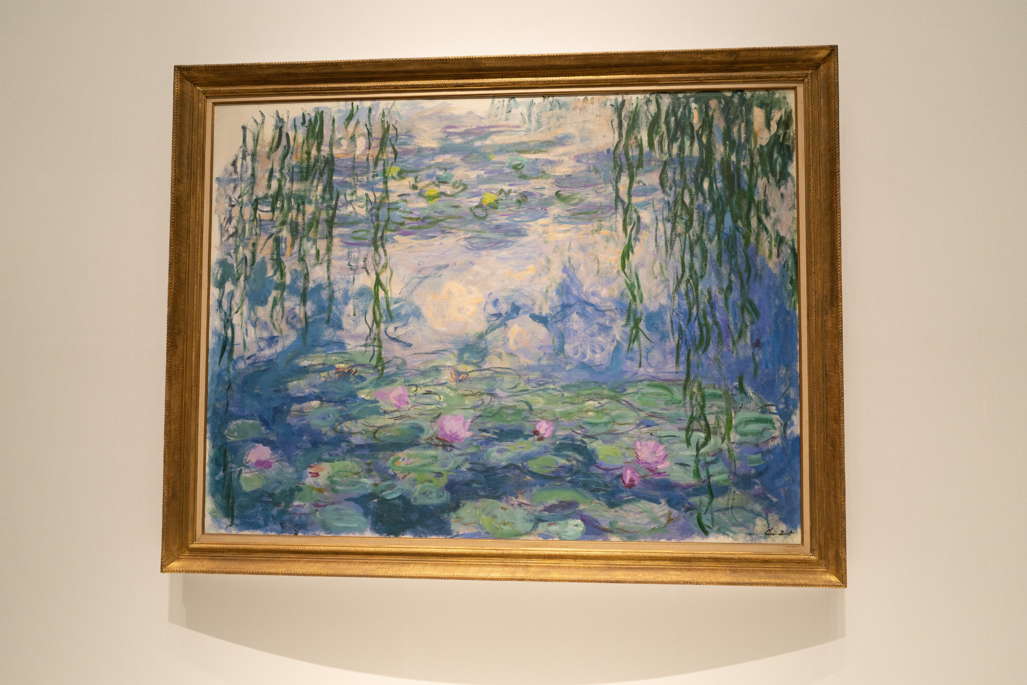 Framed Monet painting showing a pond of emerald and violet lily pads, pink blossoms, and willow branches cascading from the top against a luminous blue background.