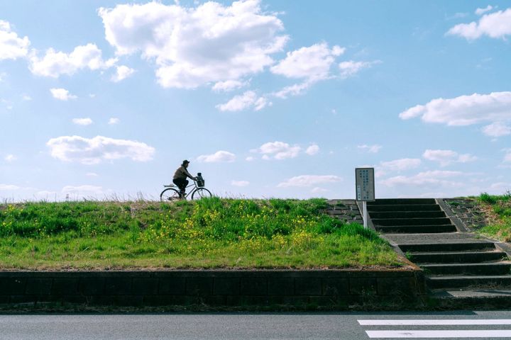晴朗春日河堤，一位骑行者剪影从绿坡掠过，象征人生旅程与日常片刻 | Photo © Philo Li