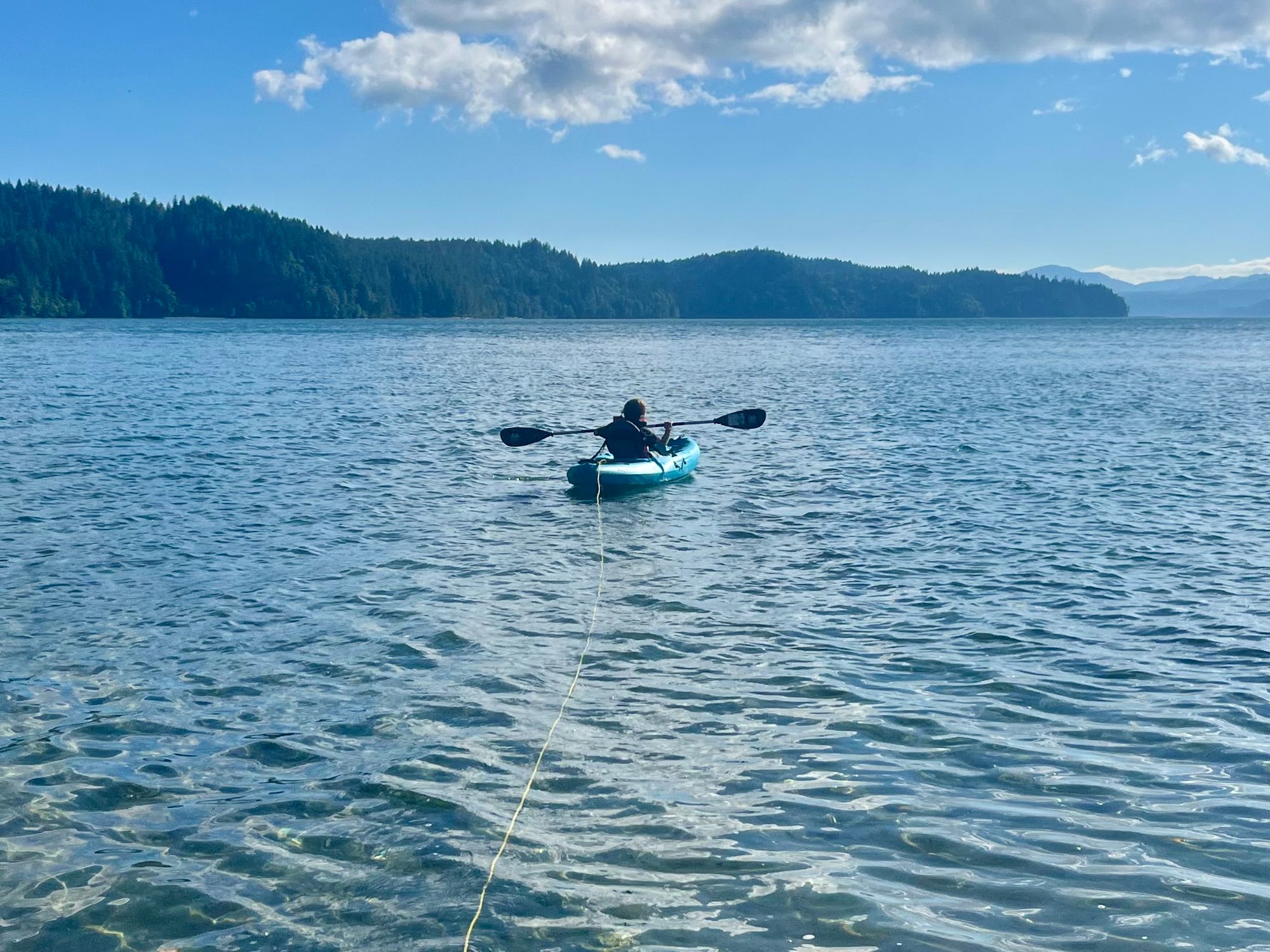 A child is in a nine-foot kayak, floating in shallow water. The kayak is tethered on a long run of rope to a buoy.