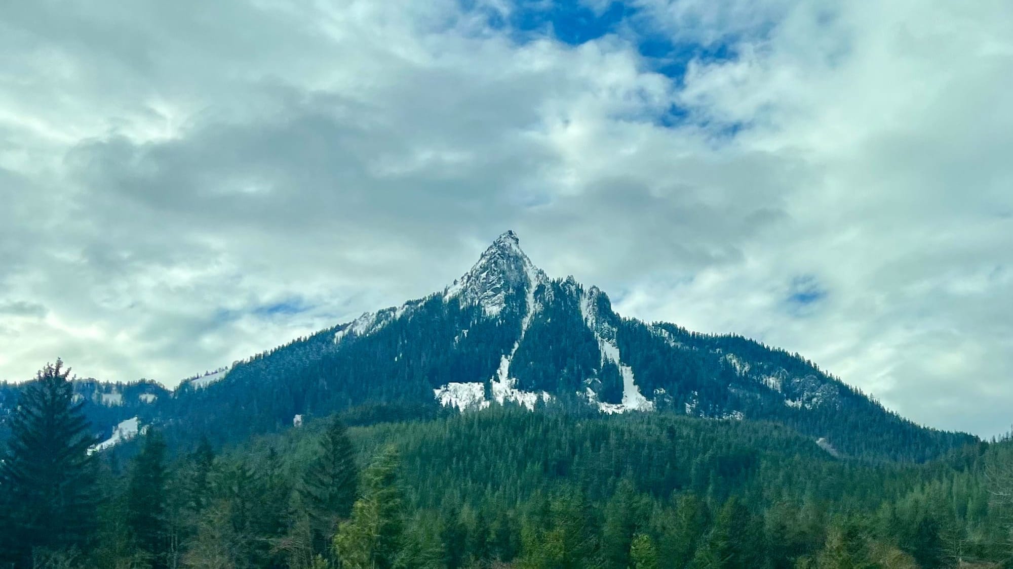 A particularly vertical mountain peak behind some trees and in front of a cloudy February morning sky.