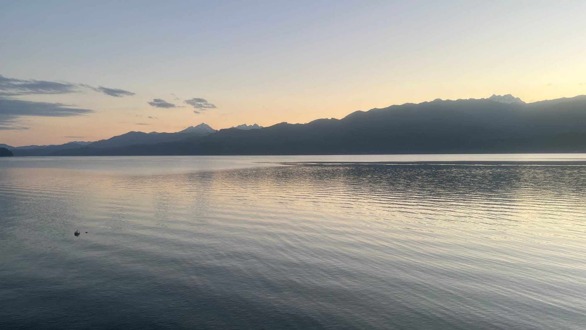 A hazy sunset, hot as hell, behind the mountains. A peachy sky with scattered clouds is reflected in water.