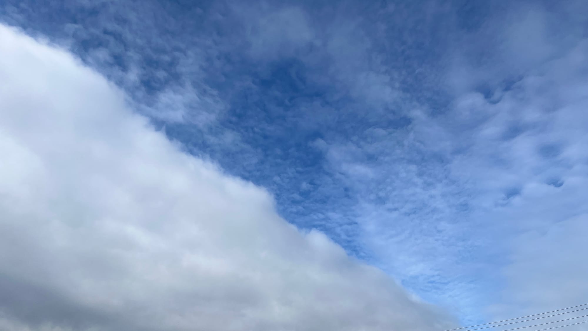 A blue sky with fluffy light clouds to the right and a solid mass of thicker rainclouds to the left. Some power lines are visible in the lower-right corner of the image.