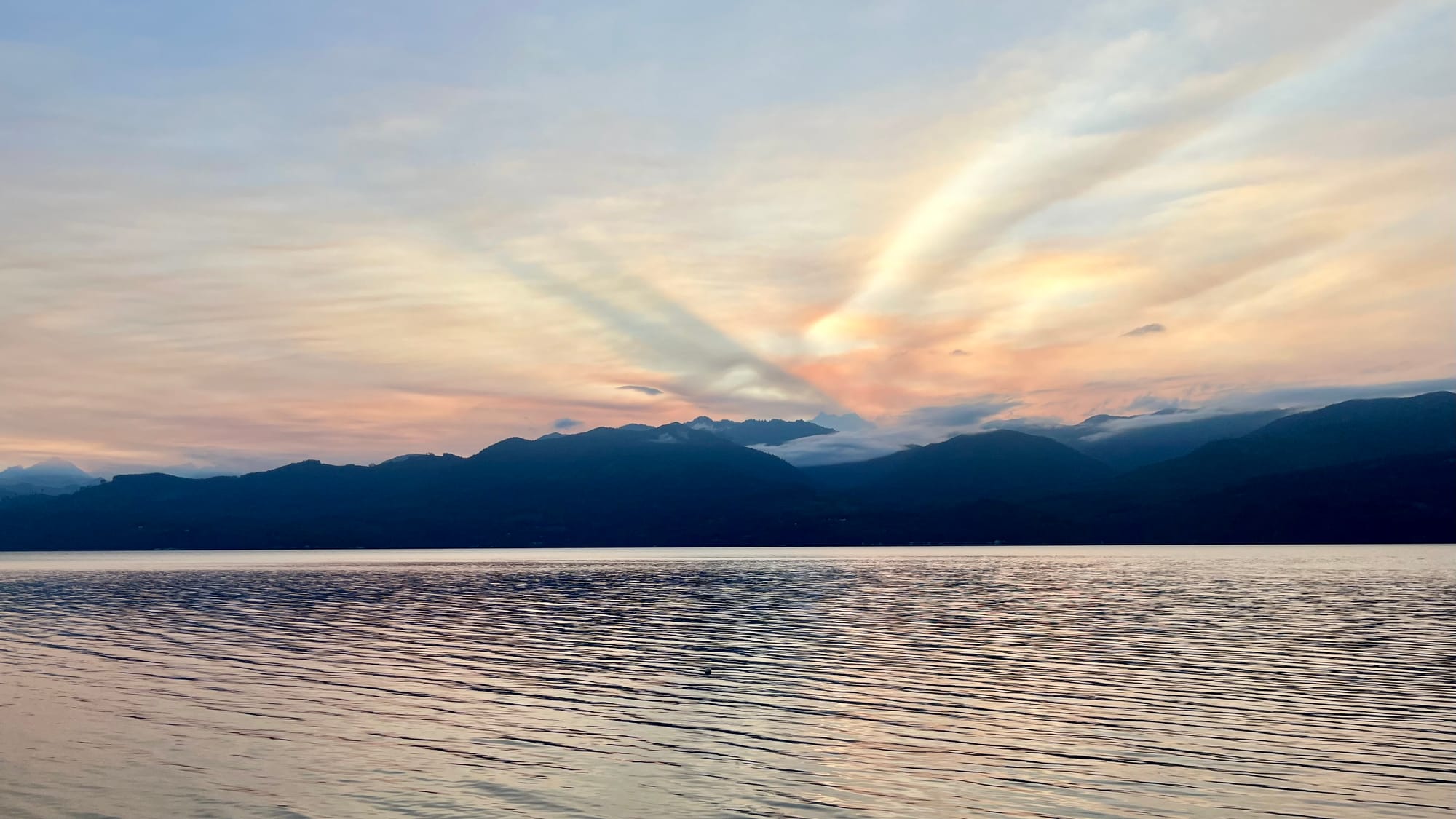 Sunset behind some wispy clouds. It’s a view of some mountains across a body of water.