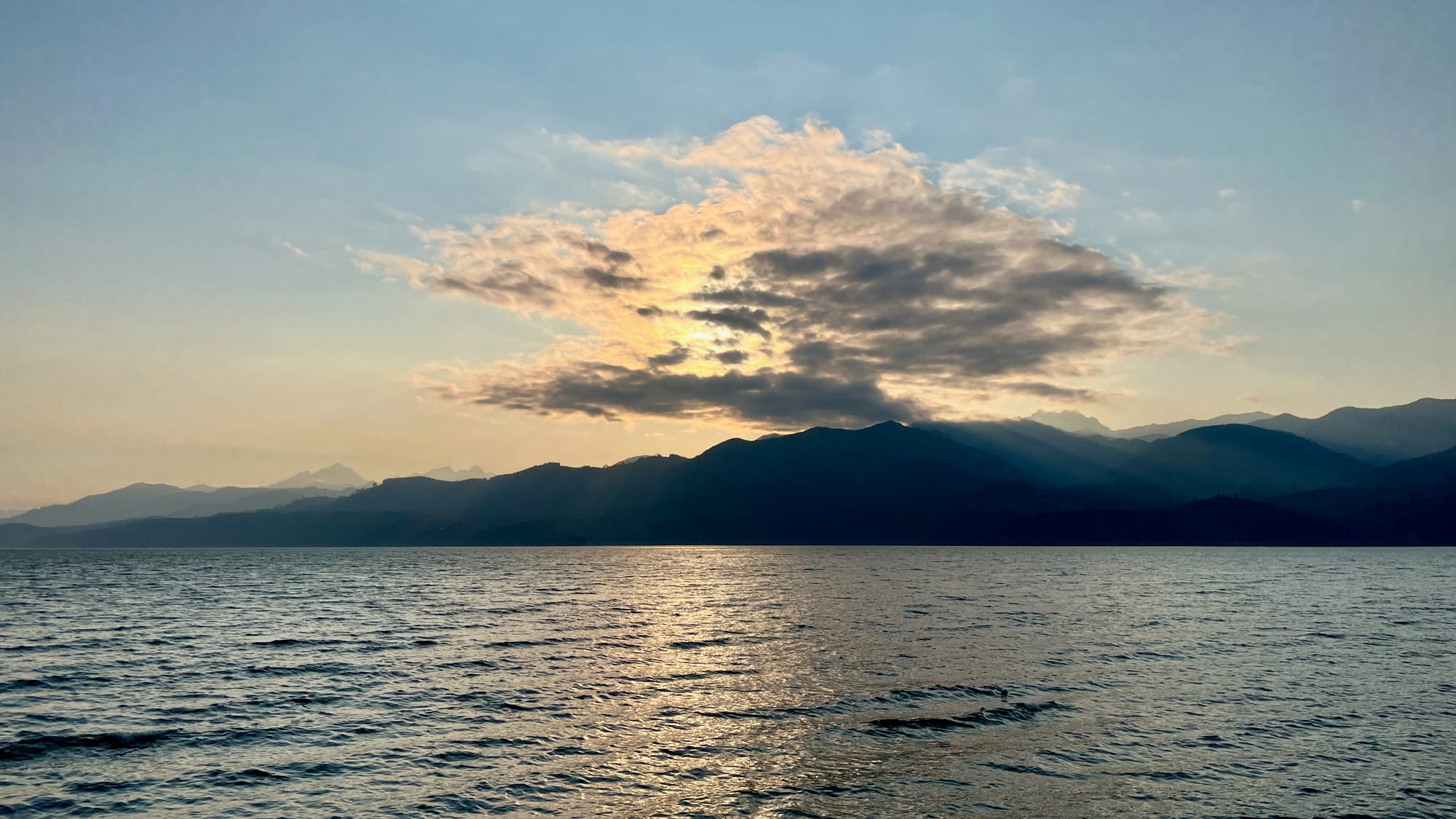 Another sunset behind the mountains, obscured by some patchy clouds, and reflected in a cool blue body of water.