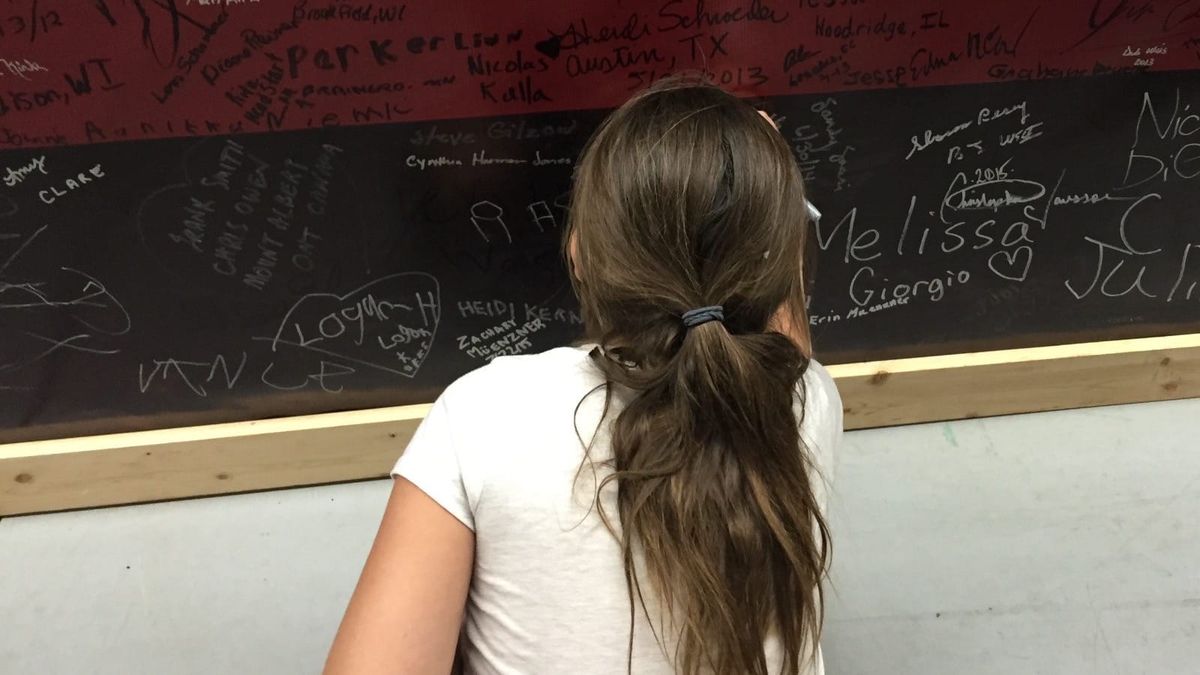 A girl with a ponytail stands facing a red-and-black wall covered with handwritten signatures. (Photo by Scott P. Richert)
