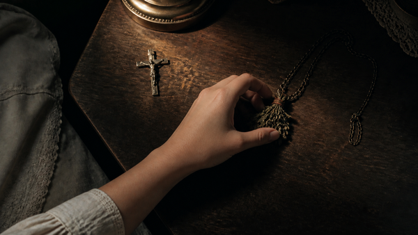 A young woman’s hand reaches past a silver crucifix to grasp an herbal charm on a dark bedside table. (AI-generated image)
