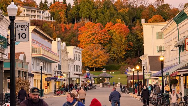 Autumn foliage rises above the storefronts on Mackinac Island’s Main Street, as people walk and cycle beneath glowing streetlamps. (Photo by Scott P. Richert)