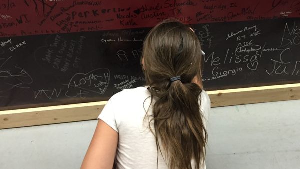 A girl with a ponytail stands facing a red-and-black wall covered with handwritten signatures. (Photo by Scott P. Richert)