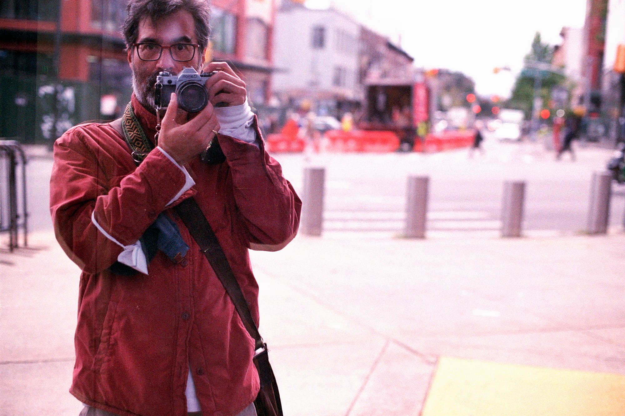 35mm color photo by Greg Pak - a photo of himself taken in a mirrored window of the Barclay Center. He's wearing a red jacket, holding a Nikon FE camera. Brooklyn streets behind him