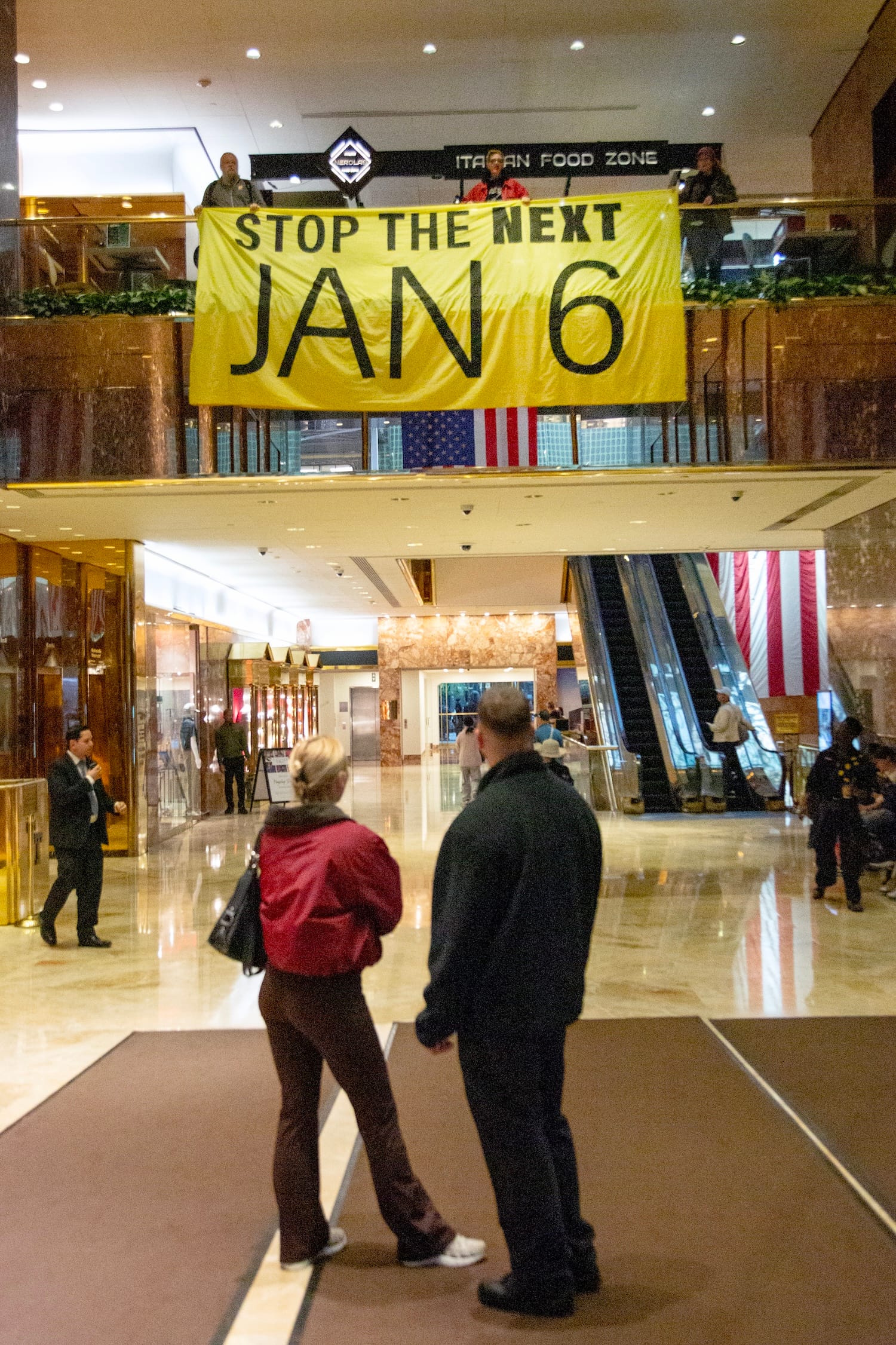 A couple of Trump Tower visitors look up at activists hanging a "STOP THE NEXT JAN 6" banner from the balcony.