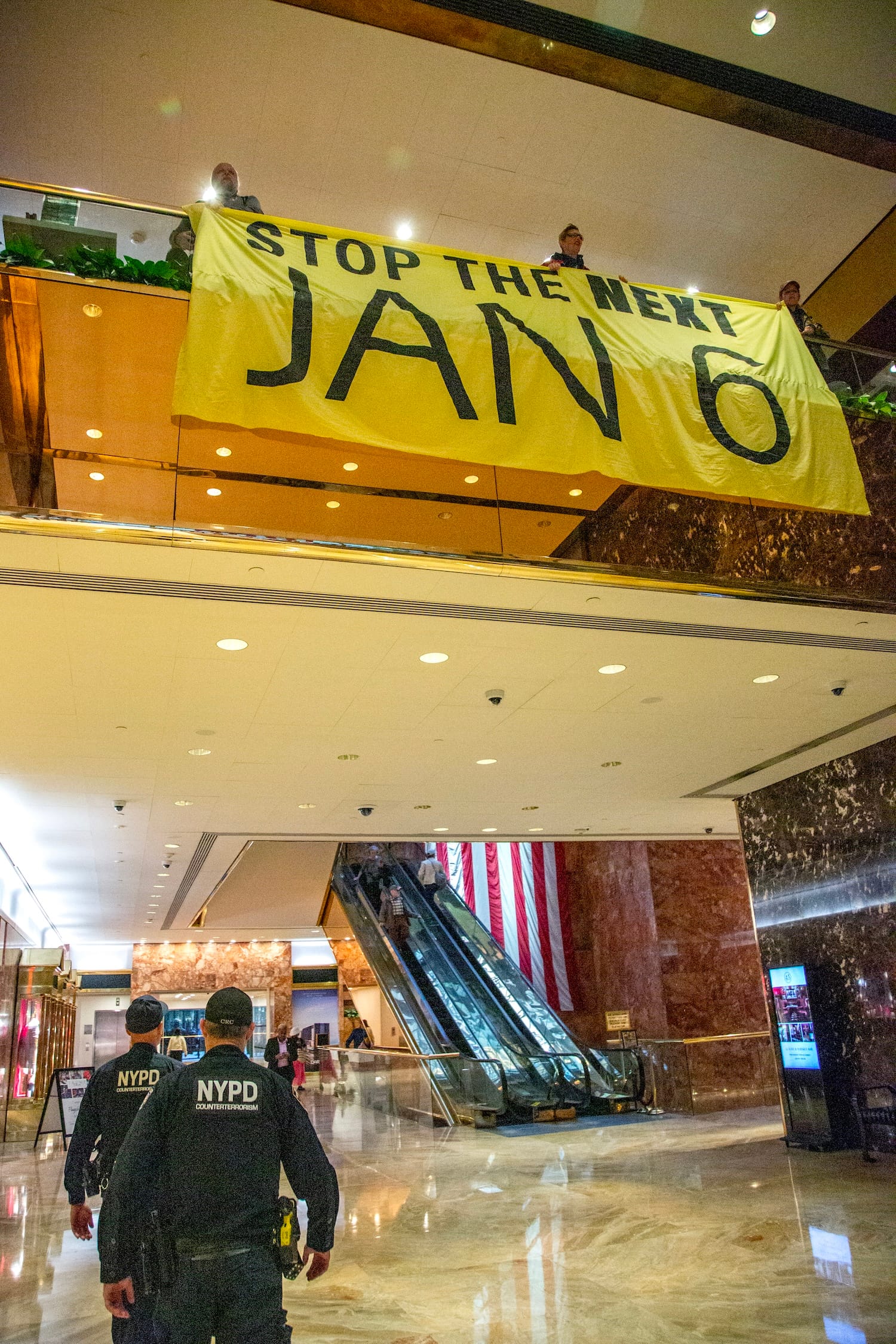 Police walk towards the escalators in Trump Tower; up above, a trio of activists hang a banner from the balcony reading "STOP THE NEXT JAN 6"