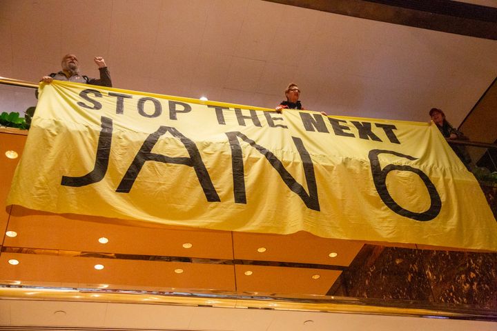 Activists chant, one with fists raised, over a banner reading "STOP THE NEXT JAN 6" on a Trump Tower balcony.