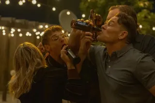 The cast of Peacemaker, Jennifer Holland, Freddie Stroma, and John Cena, drinking beer with their arms interlinked.