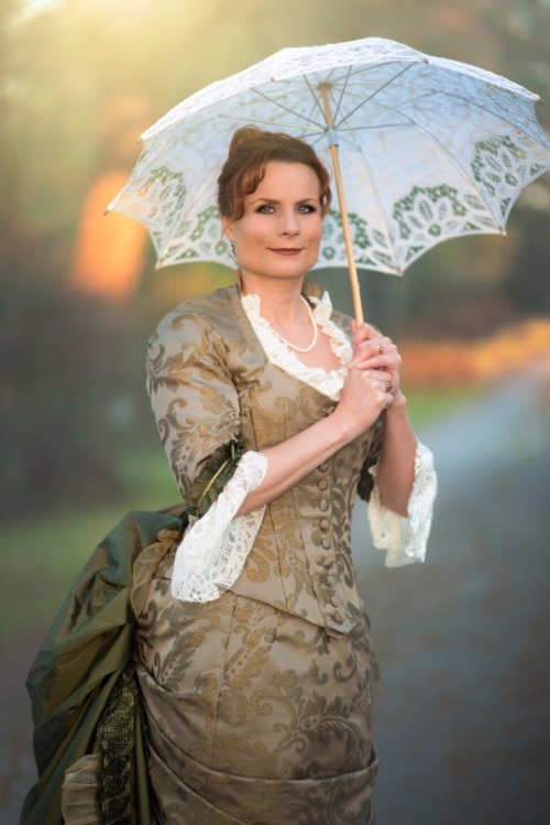  Teresa dressed in a Victorian costume of golden-green brocade, with lace at cuffs and collar, an enormous bustle, and a white lace parasol. 