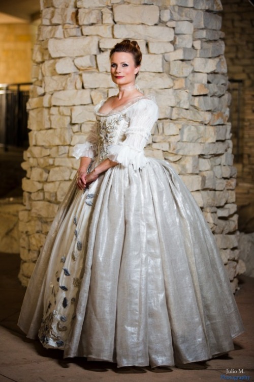  Teresa wearing the wedding dress from  Outlander —a pearl silk gown with embroidered metallic leaves all down the bodice and a very large and sweeping skirt—posing in front of a stone wall. 
