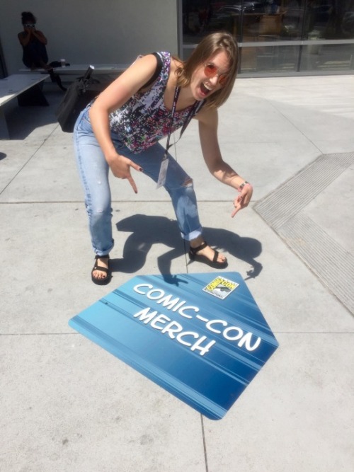  Flourish stands outside the San Diego Convention Center and points at a sign on the asphalt reading COMIC-CON MERCH. 