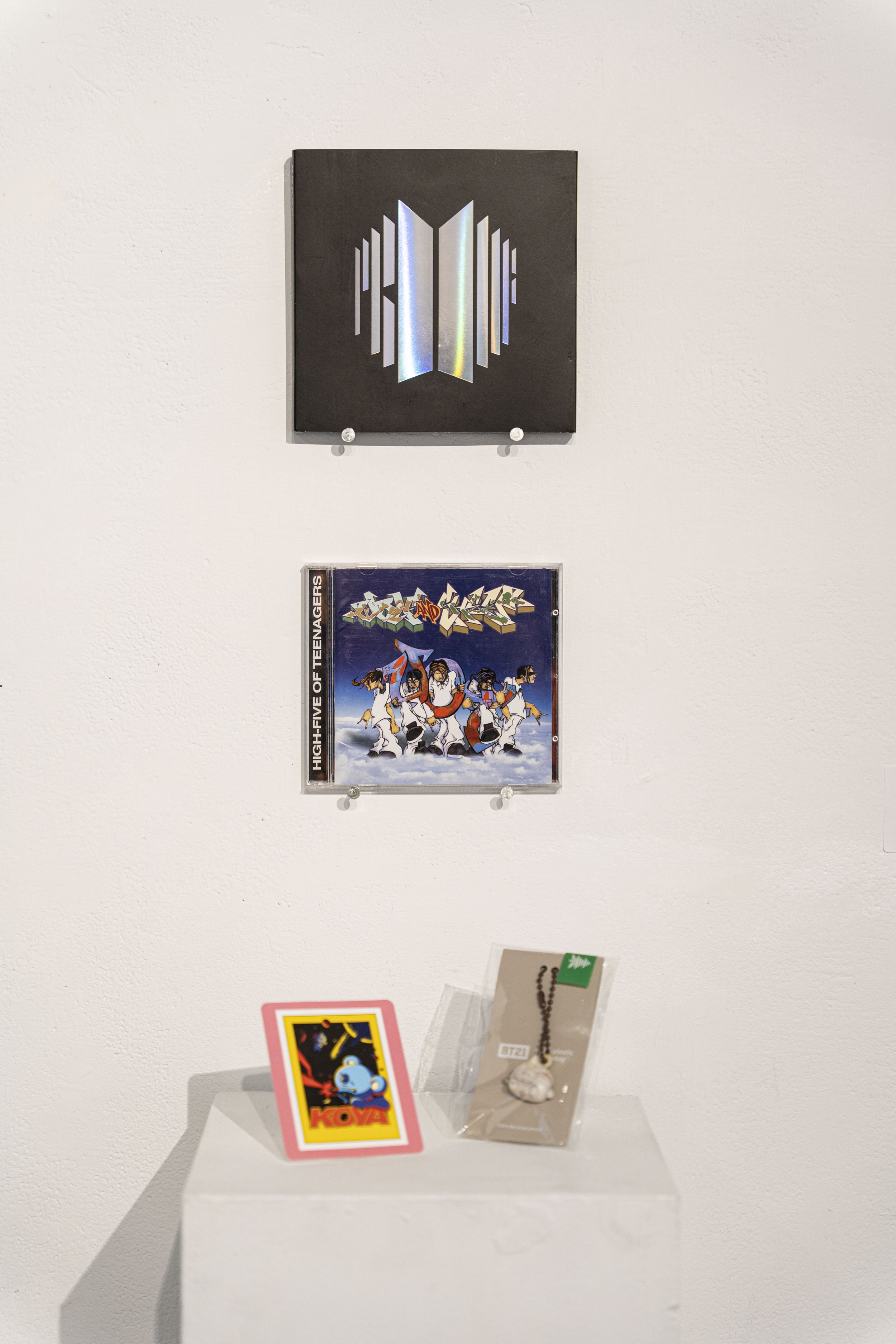 Photograph of K-pop objects against a white wall: An album in black and silver, a CD case with illustrated band members, and two objects (one card, one jewelry) on a pedastal.