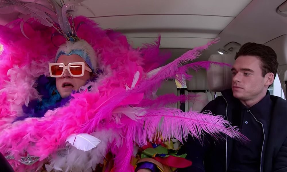 Photograph of Taron Egerton in an elaborate Elton John costume featuring many pink feathers, big sunglasses, and a headdress, sitting beside Richard Madden in all black.