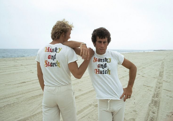 Photograph of Soul and Glasser on a beach, wearing shirts that say "Starsky and Hutch" on the front and "Husky and Starch" on the back. Glasser faces the camera, arm on Soul's shoulder. Soul faces away with his hand on Glasser's shoulder.