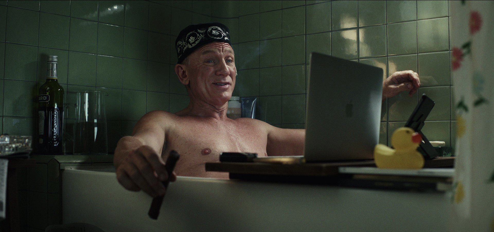 Still from Glass Onion of Benoit Blanc in the bath on a laptop, wearing a fez and smoking a cigar.