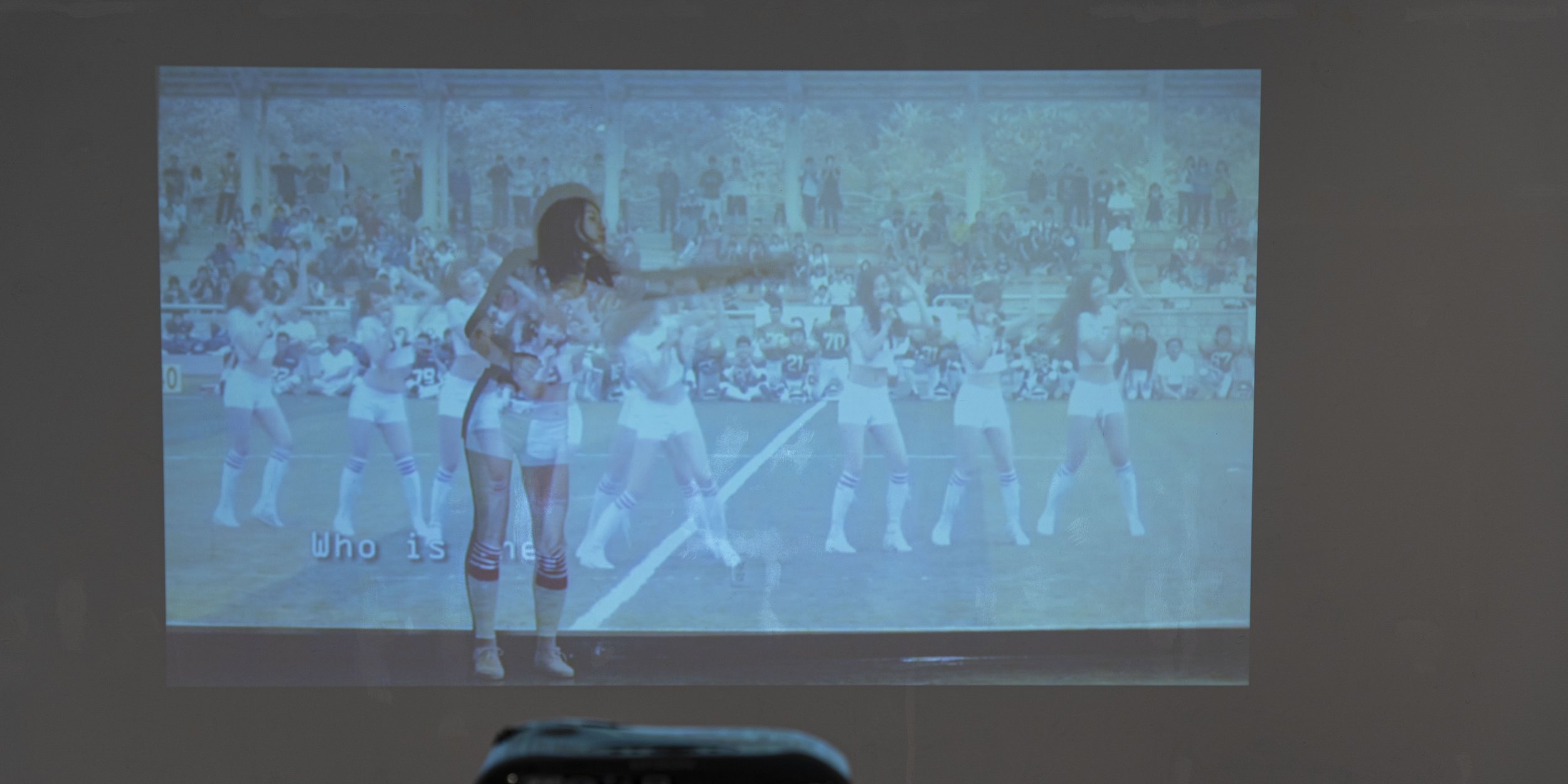 Image of Jiwon Choi in Generation Girl, in a cheerleader uniform in front of video footage of cheerleaders.