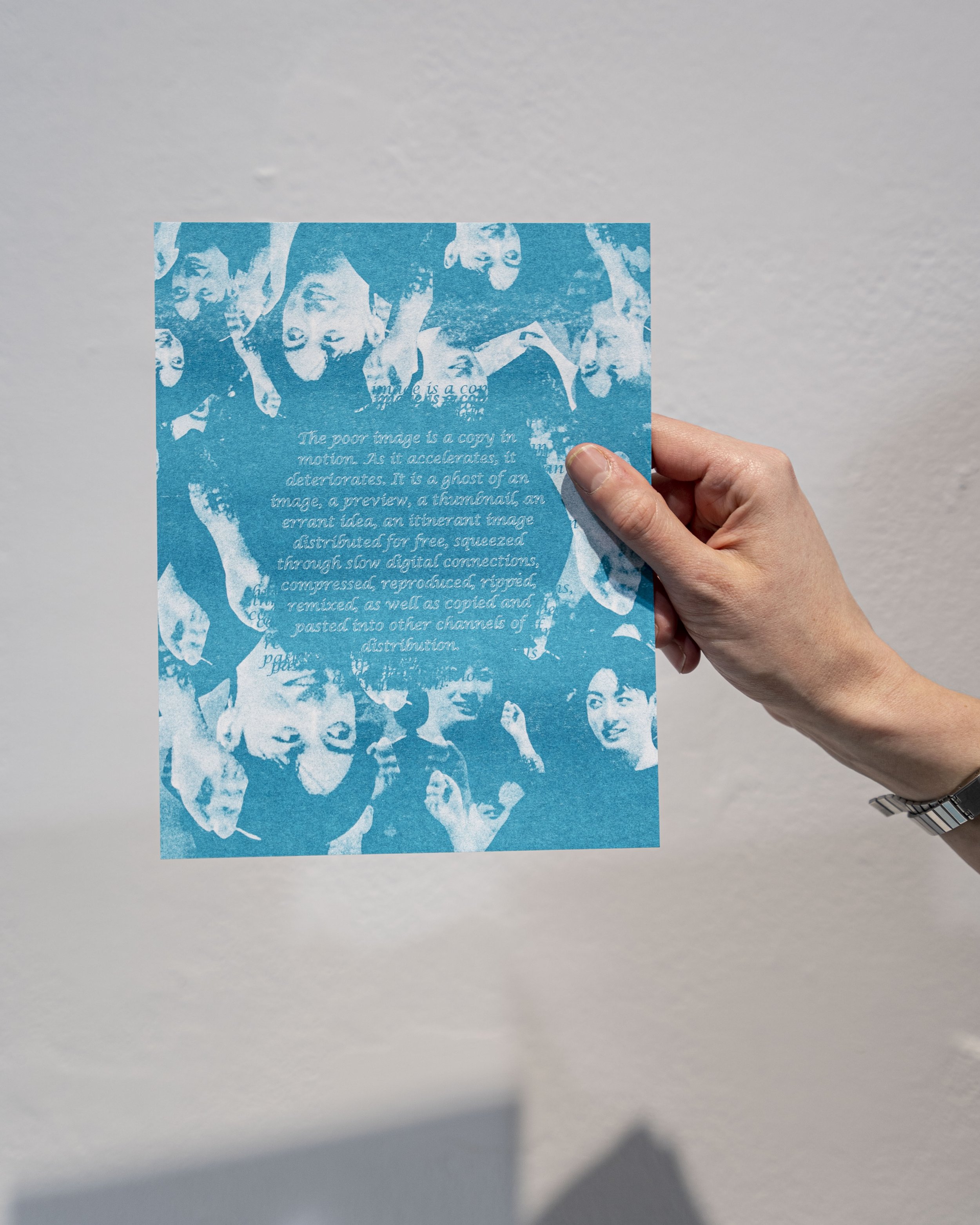 Hand holding a risograph image in washed out blue and white tones, featuring text in the center and images of K-pop artists around the edges.