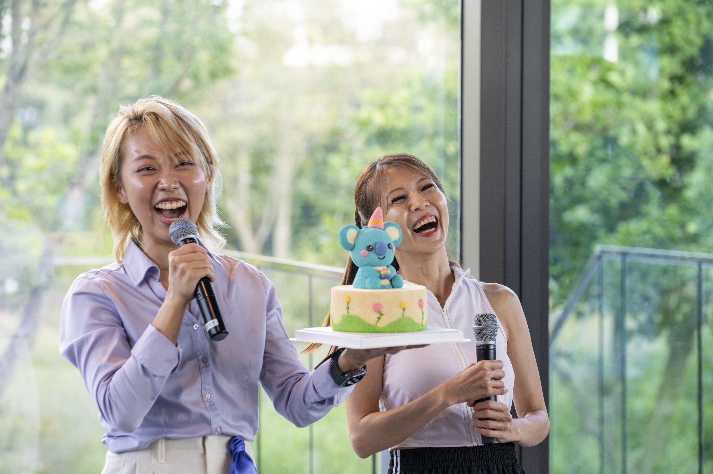 Image of two people holding mics and smiling wildly, one of them holding a cake with a small blue koala sitting on top.