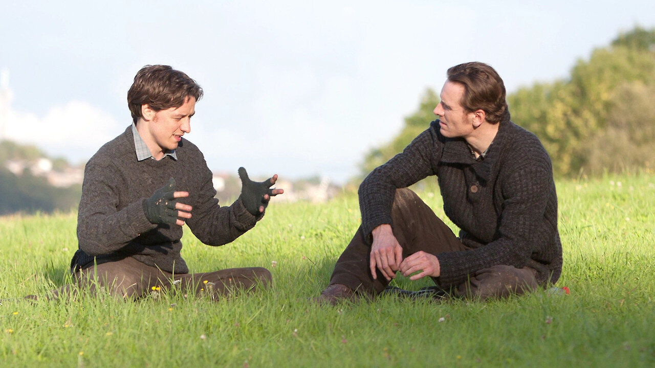 James McAvoy and Michael Fassbender sitting on the grass talking.