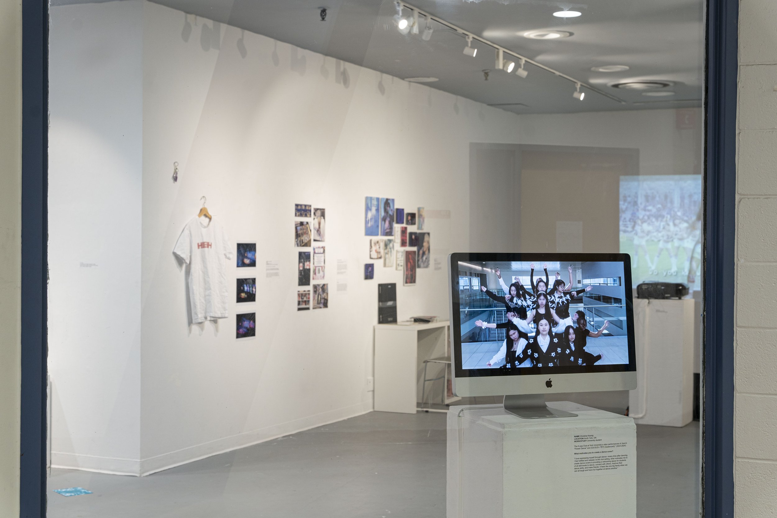 Photograph of the gallery with a computer in the foreground showing a dance performance from a dozen individuals in black and white outfits.