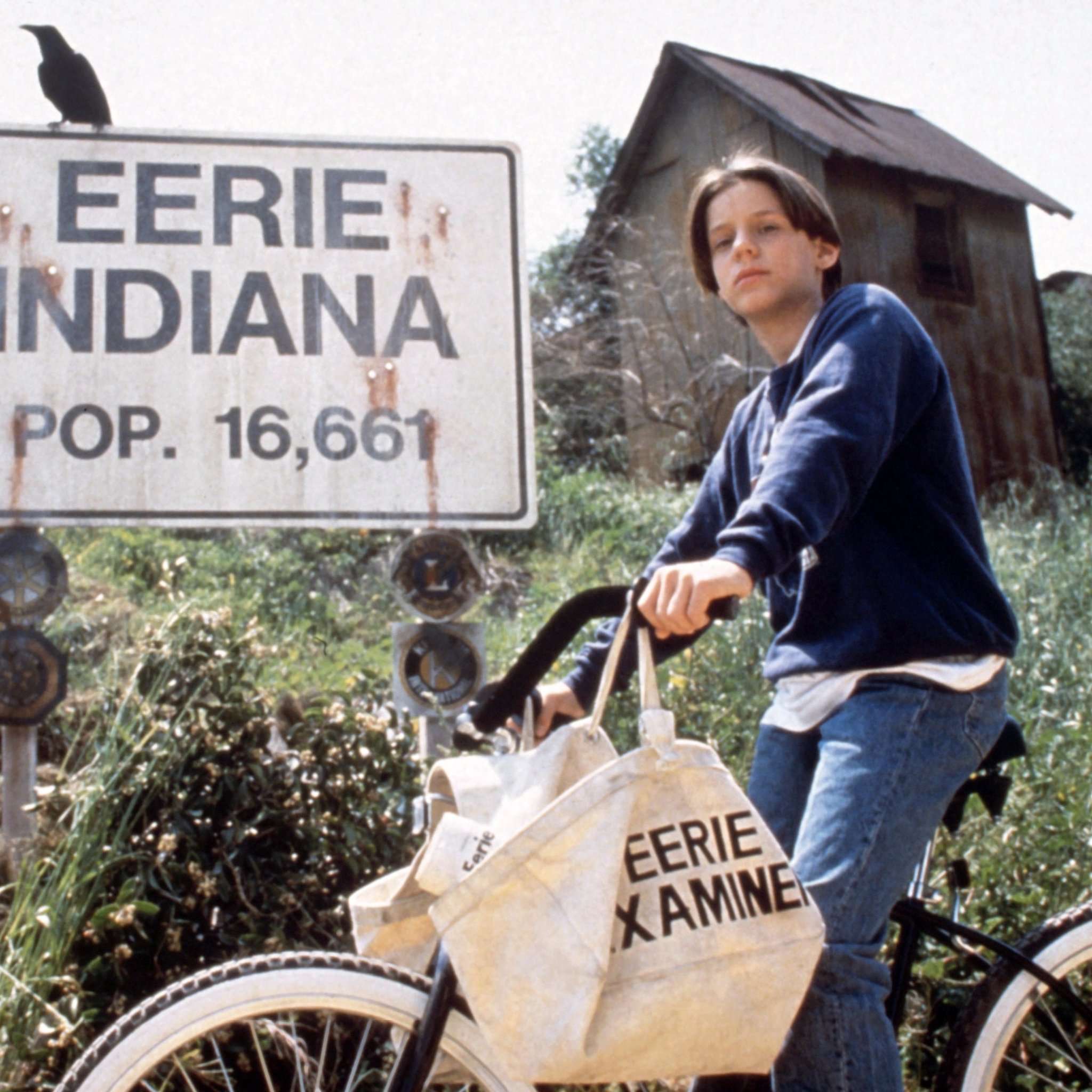Still image of Marshall from Eerie, Indiana, on a bike and standing in front of a sign reading EERIE INDIANA POP. 16,661