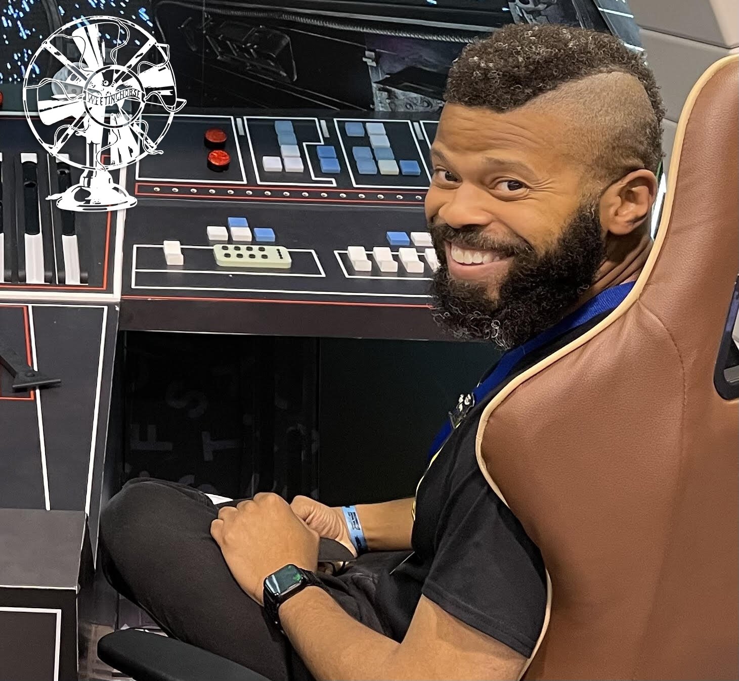 Photograph of Justin sitting in front of a control panel, grinning at the camera. 