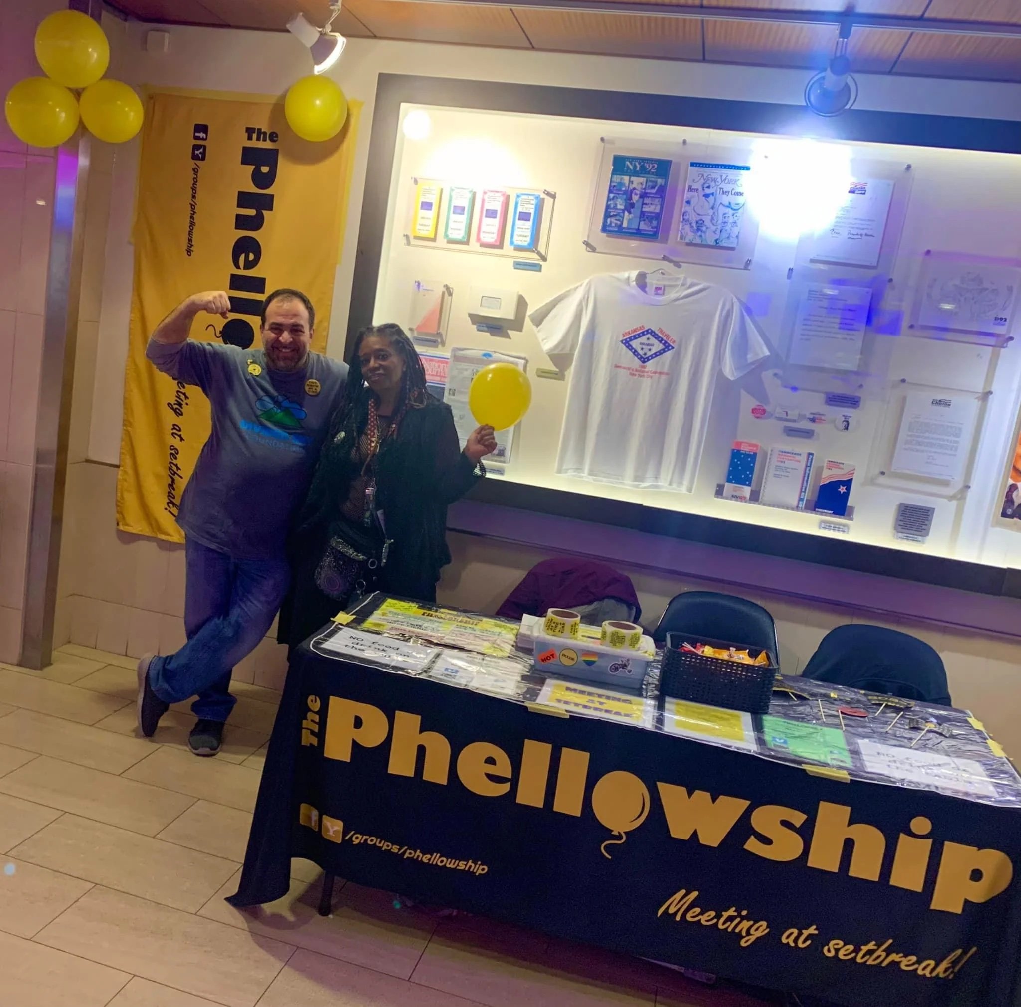 image of two people with their arms around each other beside a "Phellowship" table with a black and yellow sign.