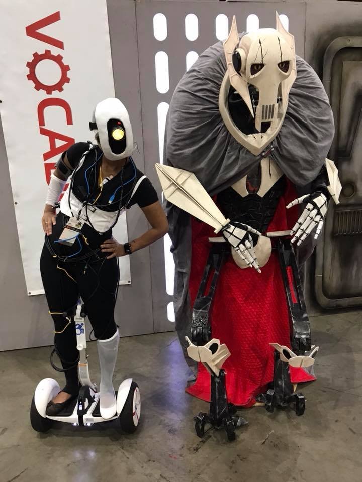 Photograph of two figures cosplaying. One wears black and white with a white object covering their face, and stands on a Segway. The other has an elaborate headpiece, gloves, and leg pieces and holds a sword.
