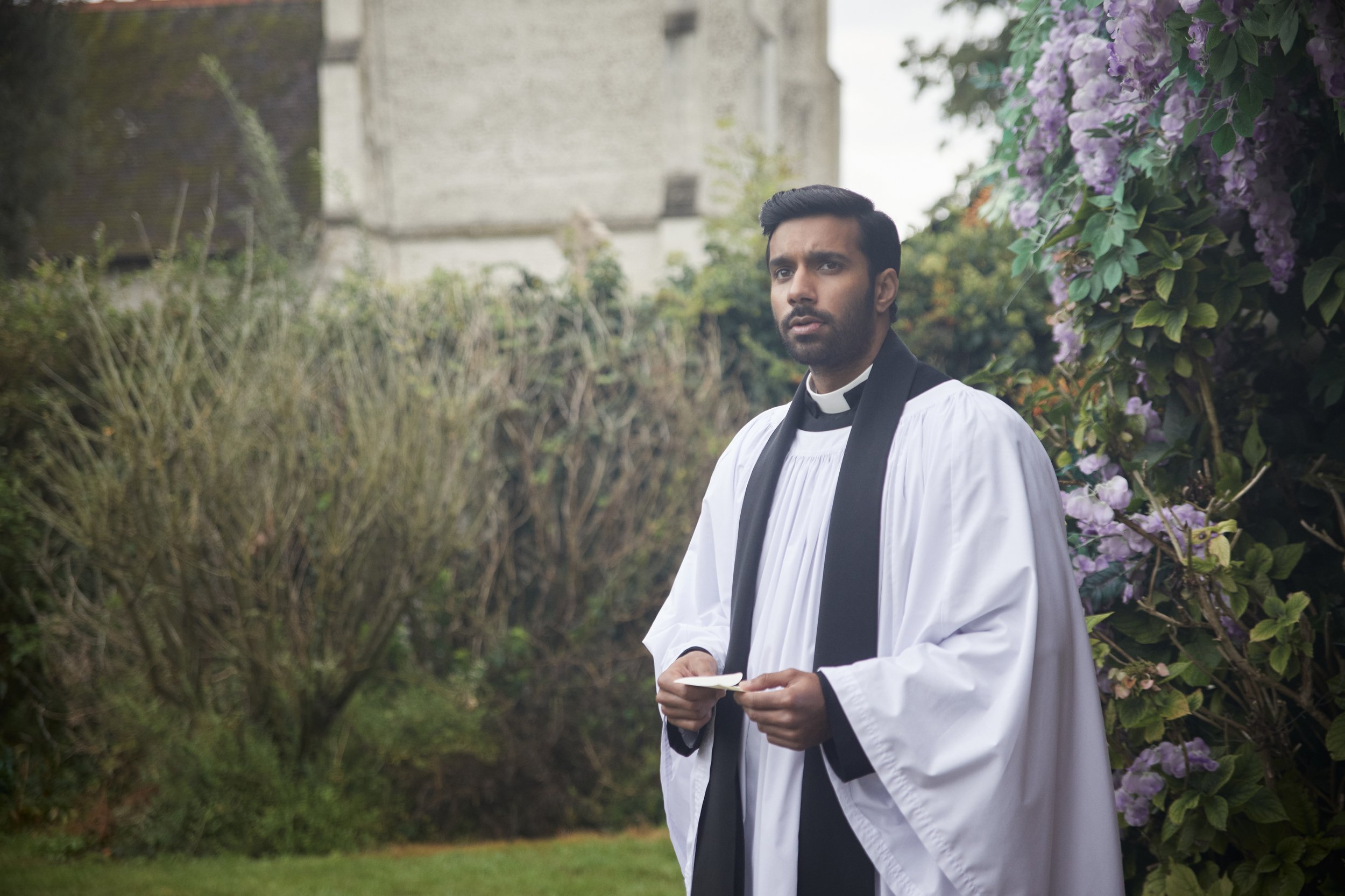 Promotional still of Rishi Nair in clerical garb standing in front of some bushes and purple flowers.